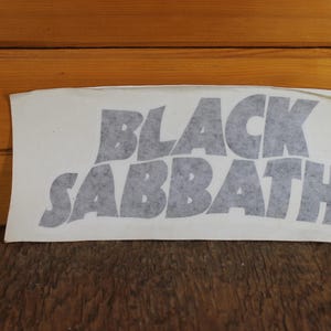 Puede incluir: Adhesivo rectangular con la inscripción "BLACK SABBATH" en letras grises estilizadas. El adhesivo está sobre un fondo blanco, con una superficie de madera en el fondo.