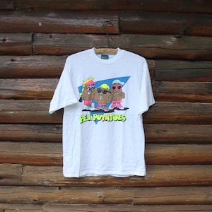 Puede incluir: Camiseta blanca con un dibujo animado de tres patatas antropomórficas con gafas de sol y sombreros, con el texto "Peel Potatoes" en verde. La camiseta está colgada sobre un fondo de madera rústica.