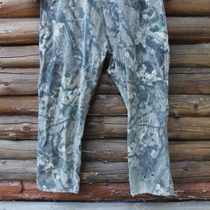 May include: Camouflage trousers in green, grey, and brown hues, set against a rustic wooden backdrop. The trousers feature a classic five-pocket design and show signs of wear, with a few small holes near the hem.