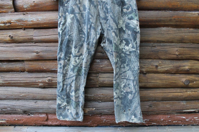 May include: Camouflage trousers in green, grey, and brown hues, set against a rustic wooden backdrop. The trousers feature a classic five-pocket design and show signs of wear, with a few small holes near the hem.