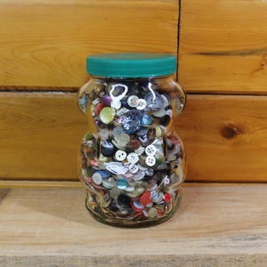 Può includere: Un barattolo di vetro trasparente a forma di orso, pieno di bottoni colorati. Il barattolo ha un coperchio verde. I bottoni sono di varie dimensioni, forme e colori, tra cui rosso, blu, bianco e giallo.