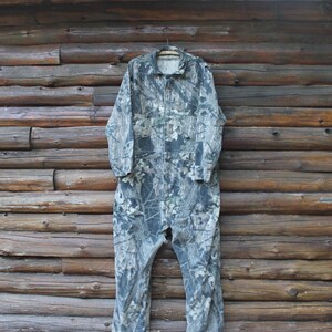 May include: A camouflage jumpsuit with a collared neckline and long sleeves hangs on a wooden hanger. The jumpsuit features a zip closure and pockets. The fabric has a pattern of green, brown, and grey, resembling a forest.