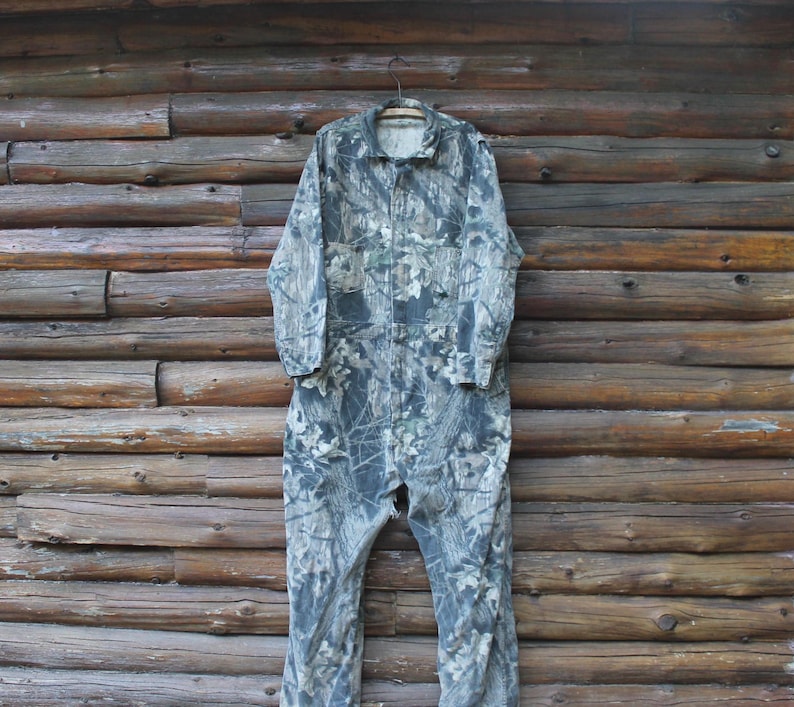 May include: A camouflage jumpsuit with a collared neckline and long sleeves hangs on a wooden hanger. The jumpsuit features a zip closure and pockets. The fabric has a pattern of green, brown, and grey, resembling a forest.