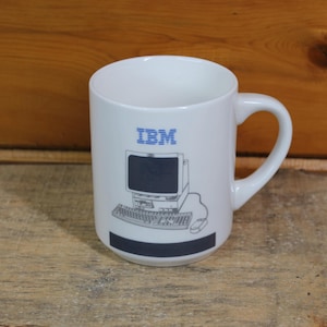 Może przedstawiać: Biały ceramiczny kubek z niebieskim logo IBM i grafiką komputera w stylu vintage. Kubek ma czarny pasek na dole i zakrzywiony uchwyt. Kubek stoi na drewnianej powierzchni.