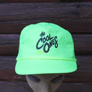 May include: A bright green cap featuring the words "the COOL ONES" in black script. The cap has a curved peak and a rope detail. The background is a wooden surface.
