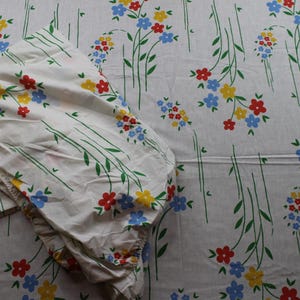 May include: A white bed sheet featuring a repeating pattern of small red, yellow, and blue flowers with green stems. The fabric is folded over on the left side, displaying the floral print across the entire surface of the textile material.