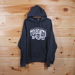 Puede incluir: Una sudadera con capucha gris oscuro con el logotipo de la Universidad de Toronto impreso en la parte delantera. El logotipo es blanco y presenta las palabras "University of Toronto" en un arco sobre un escudo con los años "1827" impresos debajo.