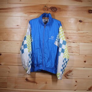 May include: A blue zip-up jacket with white and checkered sleeves. The jacket has a logo on the chest and is hanging on a metal hanger. The jacket is made of a lightweight material.