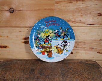 May include: A blue and white ceramic plate with a Christmas scene featuring Mickey Mouse and friends. The plate has the text "Season's Greetings" in a festive font.