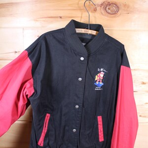 K&ouml;nnte beinhalten: Schwarze und rote Baseballjacke mit einem roten und blauen gestickten Logo eines Comic-Charakters auf der Vorderseite. Das Logo lautet "Le Hibou" und "Planet Dracula".