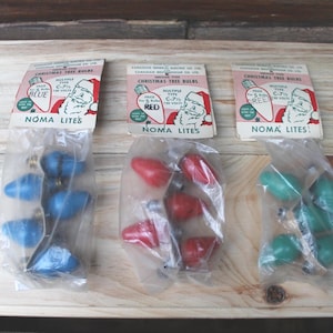Może przedstawiać: Trzy opakowania z zabytkowymi lampkami choinkowymi w kolorze niebieskim, czerwonym i zielonym. Każde opakowanie ma etykietę z ilustracją Świętego Mikołaja oraz tekstem "NOMA LITES" i "CHRISTMAS TREE BULBS". Lampki to żarówki C-7.