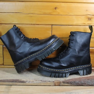 May include: A pair of black leather combat boots with chunky soles and yellow stitching. The boots feature a lace-up design and a pull tab. The boots are displayed on a wooden surface against a wood-panelled background.