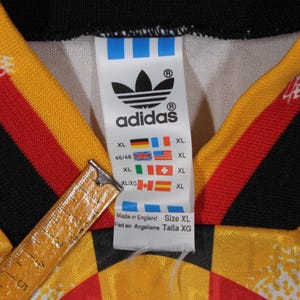 May include: A white Adidas soccer jersey with black, red, and yellow stripes. The jersey has the Adidas logo and the text "adidas" on the front. The jersey also has a size tag with the text "XL" and "Made in England".