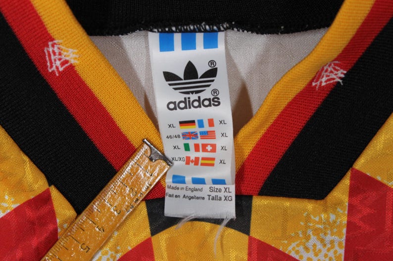 May include: A white Adidas soccer jersey with black, red, and yellow stripes. The jersey has the Adidas logo and the text "adidas" on the front. The jersey also has a size tag with the text "XL" and "Made in England".