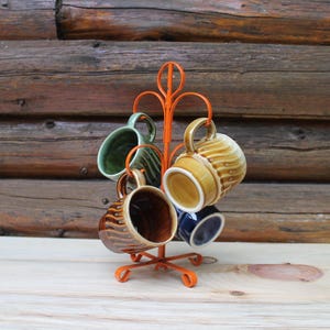 May include: An orange metal mug tree displaying six ceramic mugs. The mugs are in various colours, including green, brown, and blue, with a ribbed design. The background is a wooden wall.