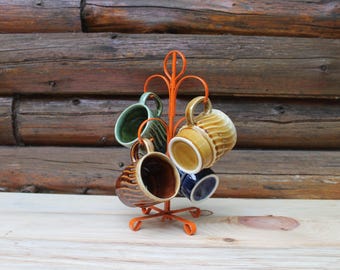 May include: An orange metal mug tree holds six ceramic mugs. The mugs are various colors, including green, brown, and blue, with a ribbed design. The background is a wooden wall.