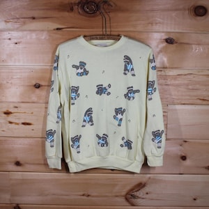 Puede incluir: Una sudadera amarilla con cuello redondo y mangas largas. La sudadera presenta un patrón repetido de osos marrones que llevan camisas y pantalones a rayas azules y blancas. Los osos están en varias poses, incluyendo bailar y saltar.