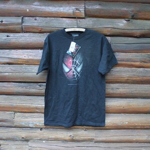 May include: A black short-sleeve t-shirt featuring a graphic of a Spider-Man mask on the chest. The mask design is split, with one side showing a red and black webbed pattern and the other side in a grayscale webbed texture. A tag is attached.