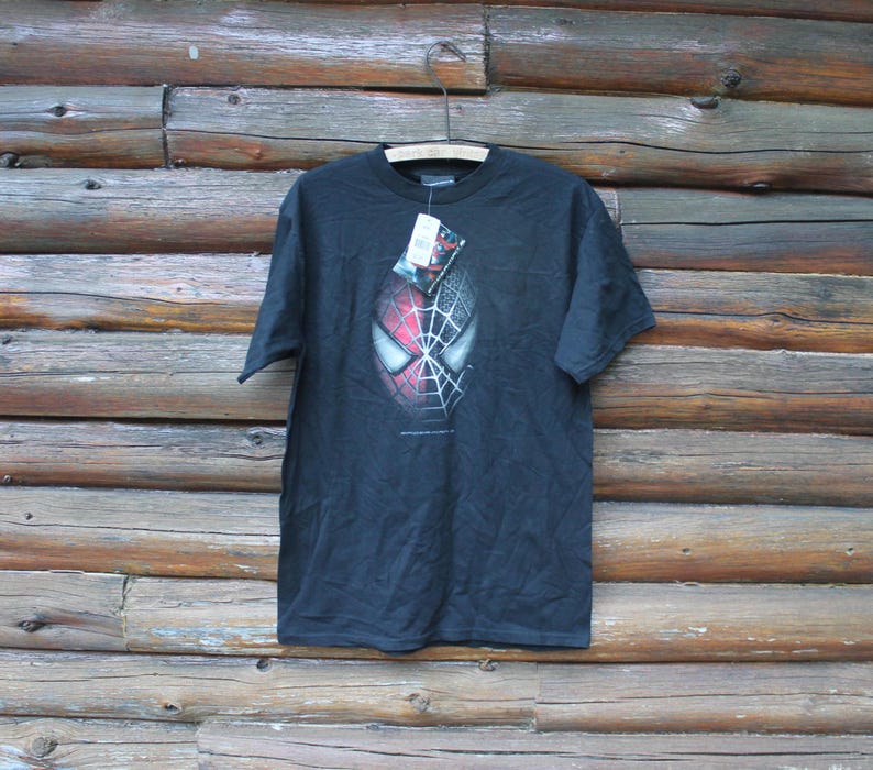 May include: A black short-sleeve t-shirt featuring a graphic of a Spider-Man mask on the chest. The mask design is split, with one side showing a red and black webbed pattern and the other side in a grayscale webbed texture. A tag is attached.