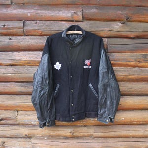 Op de afbeelding: Zwarte varsity jas met leren mouwen. Voorzien van een witte esdoornblad patch en een rood-wit logo. De jas heeft een knoopsluiting, een zwarte kraag en twee voorzakken. De jas hangt aan een houten kleerhanger.