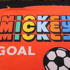 May include: Orange towel featuring the word "MICKEY" in colorful block letters, with "GOAL" in white below. A black and white soccer ball is on the bottom right. The towel is likely a sports-themed item.