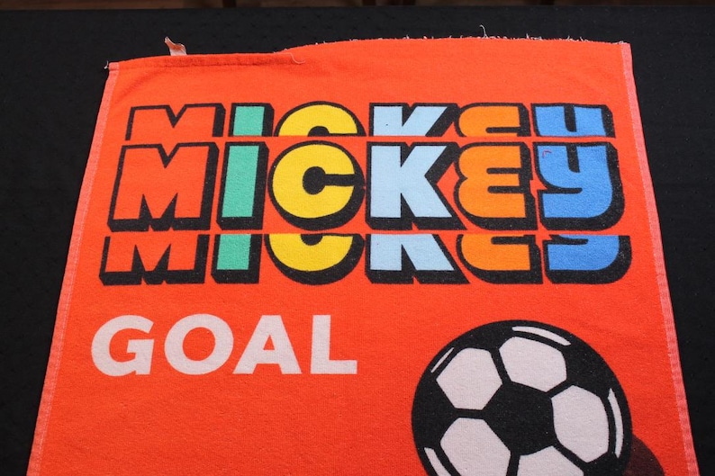 May include: Orange towel featuring the word "MICKEY" in colorful block letters, with "GOAL" in white below. A black and white soccer ball is on the bottom right. The towel is likely a sports-themed item.