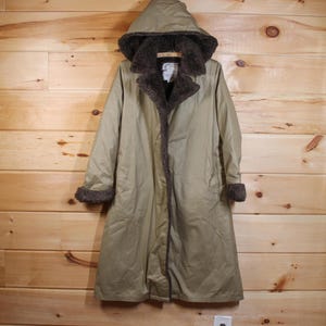 Puede incluir: Un abrigo largo verde oliva con capucha y forro de piel sintética. El abrigo tiene cierre de botones y ribete de piel en los puños y el cuello. El abrigo está colgado.