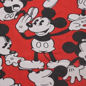 Op de afbeelding: Rode stof met een herhalend patroon van zwarte en witte Mickey Mouse-figuren. De Mickey Mouse-figuren staan in verschillende poses, waaronder zwaaien, glimlachen en verbaasd kijken.