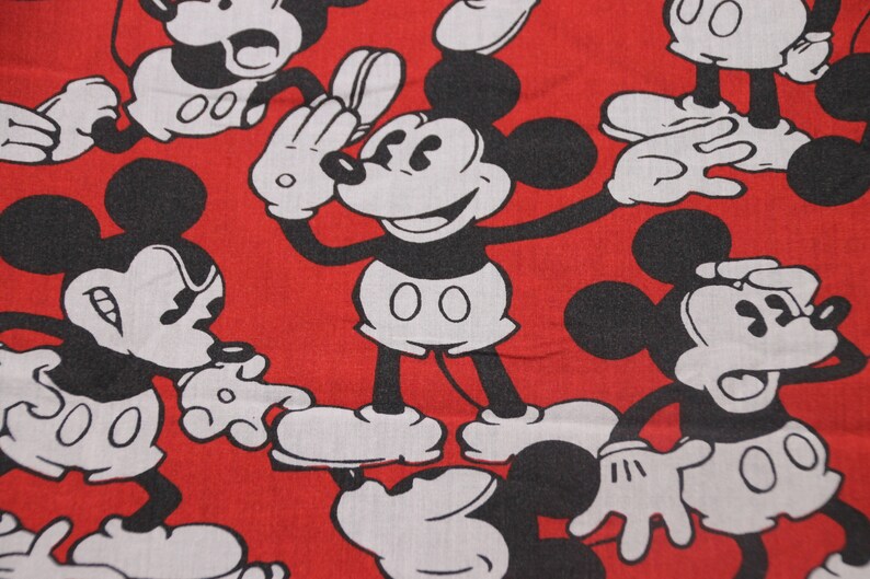 Op de afbeelding: Rode stof met een herhalend patroon van zwarte en witte Mickey Mouse-figuren. De Mickey Mouse-figuren staan in verschillende poses, waaronder zwaaien, glimlachen en verbaasd kijken.