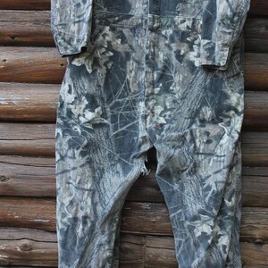 May include: Camouflage-patterned coveralls in shades of green, grey, and brown. The one-piece garment features a button-up front, long sleeves, and a relaxed fit. The fabric has a detailed print of leaves and branches.