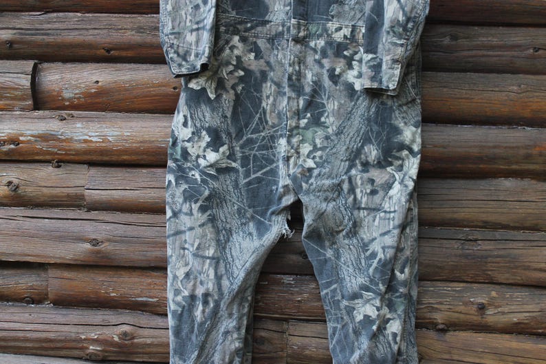 May include: Camouflage-patterned coveralls in shades of green, grey, and brown. The one-piece garment features a button-up front, long sleeves, and a relaxed fit. The fabric has a detailed print of leaves and branches.