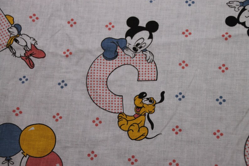 May include: A white fabric with a red and blue polka dot pattern. Mickey Mouse, Pluto, and Donald Duck are featured on the fabric. Mickey Mouse is sitting on a red and white polka dot letter C. Pluto is sitting next to the letter C. Donald Duck is in the upper left corner of the fabric.