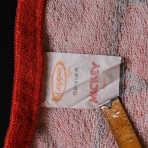 May include: A close-up of a towel with a red border and a pink and white patterned center. A tag with the text "Lepper Kids" and "Mickey" is visible. The towel measures 70 cm x 1.40 m.