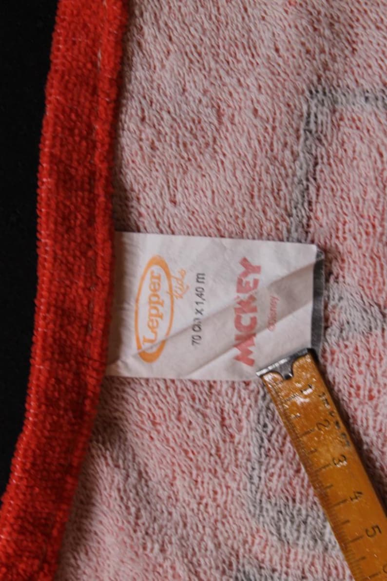 May include: A close-up of a towel with a red border and a pink and white patterned center. A tag with the text "Lepper Kids" and "Mickey" is visible. The towel measures 70 cm x 1.40 m.