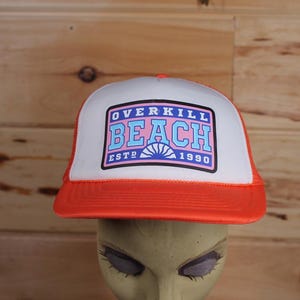 May include: A white and orange trucker hat with a blue, pink, and white logo that says "Overkill Beach Est. 1990".