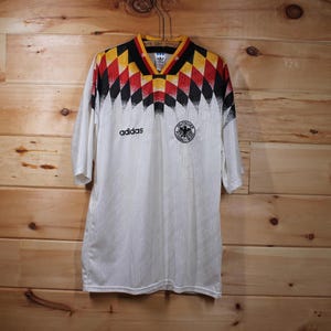 May include: White soccer jersey with black and yellow geometric pattern, Adidas logo, and the German national team crest. The jersey has a white background with a black and yellow geometric pattern on the shoulders and sleeves.