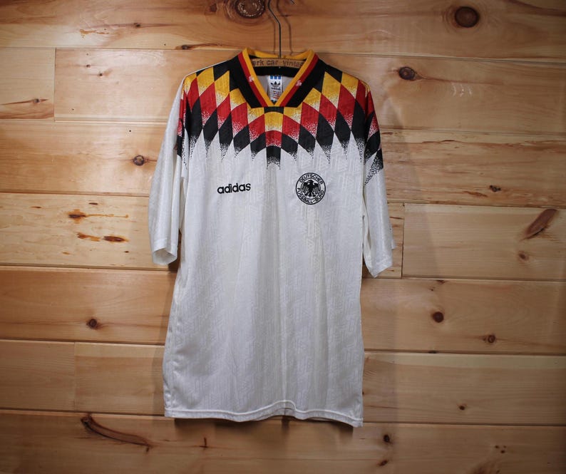 May include: White soccer jersey with black and yellow geometric pattern, Adidas logo, and the German national team crest. The jersey has a white background with a black and yellow geometric pattern on the shoulders and sleeves.