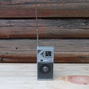 May include: A vintage grey portable radio with a telescopic aerial. The radio features a speaker grille, AM/FM dial, and the brand name "Prosonic". The radio is rectangular and rests on a wooden surface.