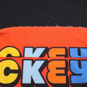 May include: Close-up of an orange fabric with the word "MICKEY" in large, colorful letters. The letters are outlined in black and filled with yellow, blue, green, and orange. The background is black.