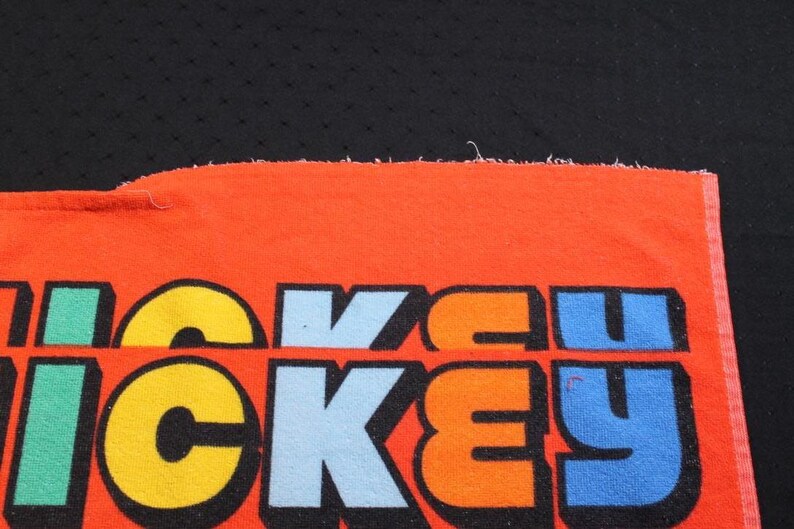 May include: Close-up of an orange fabric with the word "MICKEY" in large, colorful letters. The letters are outlined in black and filled with yellow, blue, green, and orange. The background is black.
