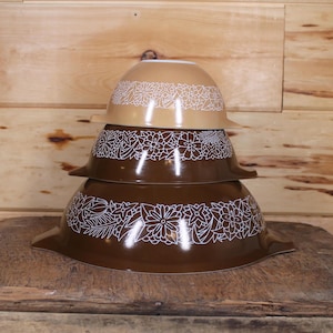 Puede incluir: Tres cuencos de mezcla Pyrex vintage apilados sobre una superficie de madera. Los cuencos son marrones y beige con un diseño floral blanco alrededor del borde. El fondo es una pared de madera.