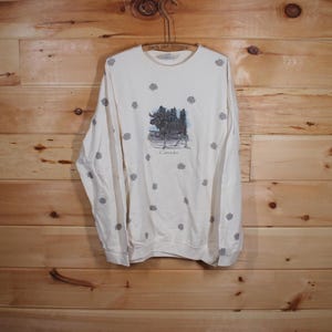 Op de afbeelding: Een wit longsleeve sweatshirt met een eland en bomen afbeelding en het woord "Canada" gedrukt op de voorkant. Het sweatshirt heeft een klein, grijs sneeuwvlokkenpatroon.