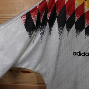 May include: White soccer jersey with a black, red, and yellow geometric pattern on the right shoulder. The Adidas logo is printed in black on the front of the jersey. The jersey has a small black and white logo on the left sleeve.