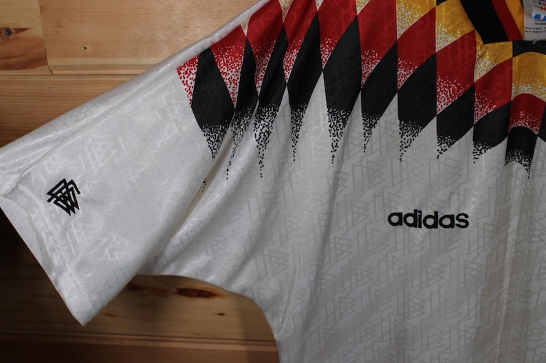 May include: White soccer jersey with a black, red, and yellow geometric pattern on the right shoulder. The Adidas logo is printed in black on the front of the jersey. The jersey has a small black and white logo on the left sleeve.