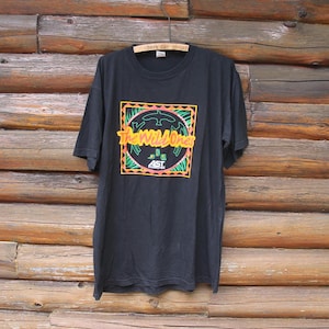 May include: Black t-shirt featuring a graphic of a circle with the text "The Wild Ones" and a logo in the centre. The circle is surrounded by a green, orange and yellow design.