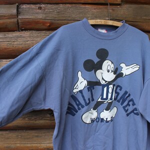 Puede incluir: Sudadera azul de manga larga con un dibujo de Mickey Mouse y el texto "WALT DISNEY WORLD". El personaje de dibujos animados es en negro, blanco y azul. La sudadera est&aacute; colgada en una percha de madera.
