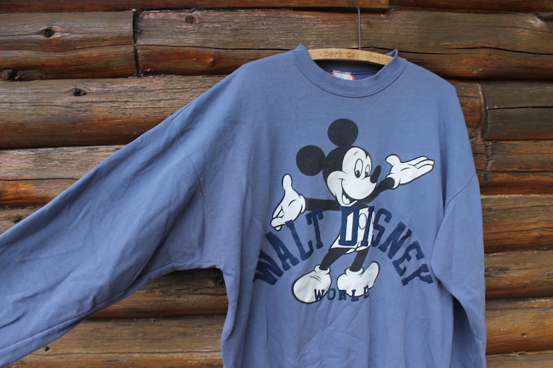 Puede incluir: Sudadera azul de manga larga con un dibujo de Mickey Mouse y el texto "WALT DISNEY WORLD". El personaje de dibujos animados es en negro, blanco y azul. La sudadera est&aacute; colgada en una percha de madera.