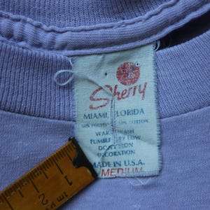 May include: A light purple t-shirt with a white fabric tag featuring the red Sherry logo, Miami, Florida text, and care instructions. A yellow measuring tape is positioned next to the tag, showing a length of approximately 2 inches for the label.