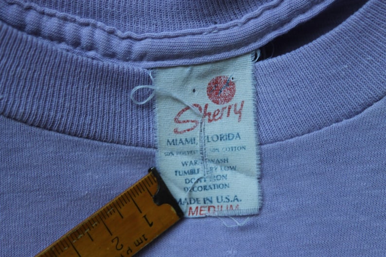May include: A light purple t-shirt with a white fabric tag featuring the red Sherry logo, Miami, Florida text, and care instructions. A yellow measuring tape is positioned next to the tag, showing a length of approximately 2 inches for the label.