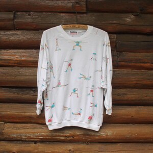 May include: White long-sleeve sweatshirt with a colorful print of stick figures doing various exercises. The text "BAZZL" is printed on the front of the shirt.